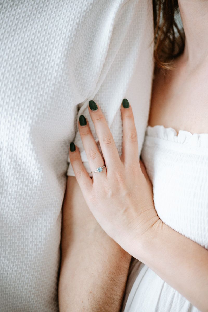 A hand with dark green nail polish rests against white fabric, wearing a delicate ring on one finger.