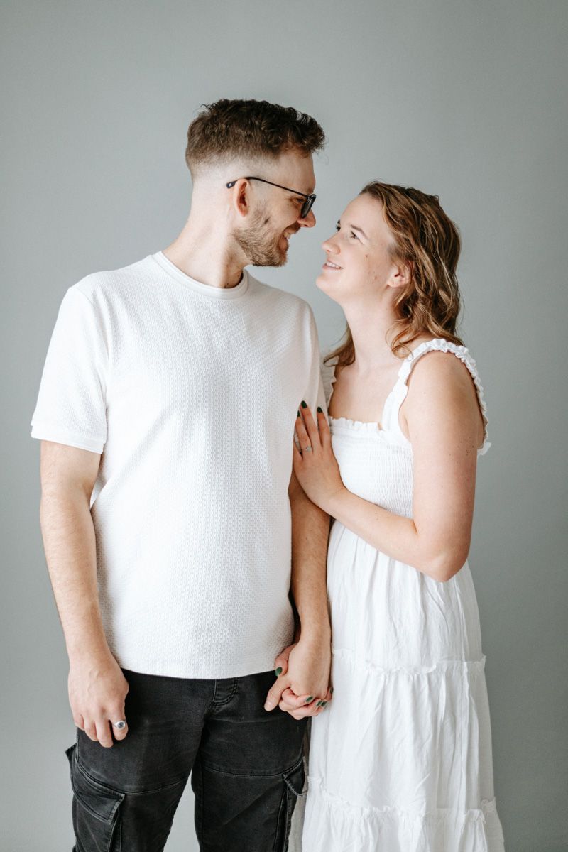 A couple in white outfits shares intimate moments while posing against a gray studio backdrop.