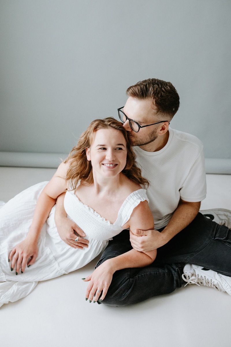 A couple in casual white attire share an intimate moment while sitting together on a light colored surface.