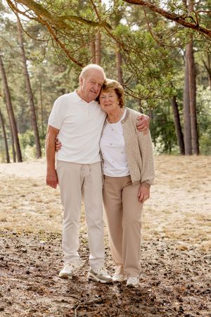 opa en oma in wit/beige kleding tijdens een familieshoot op een zandvlakte onder een naaldboom, houden elkaar vast, kijkend richting camera.