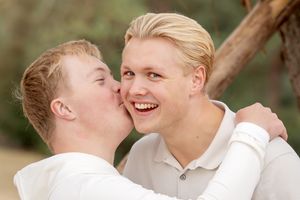 2 broers. oudste blonde jongen kijkt in camera. jongere broertje met downsyndroom geeft hem een kus op zijn wang.
