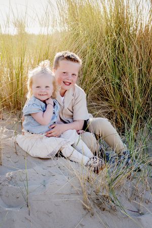 broer (5) en zus (2) zitten knuffelend tussen het hoge gras in de duinen bij ondergaande zon.