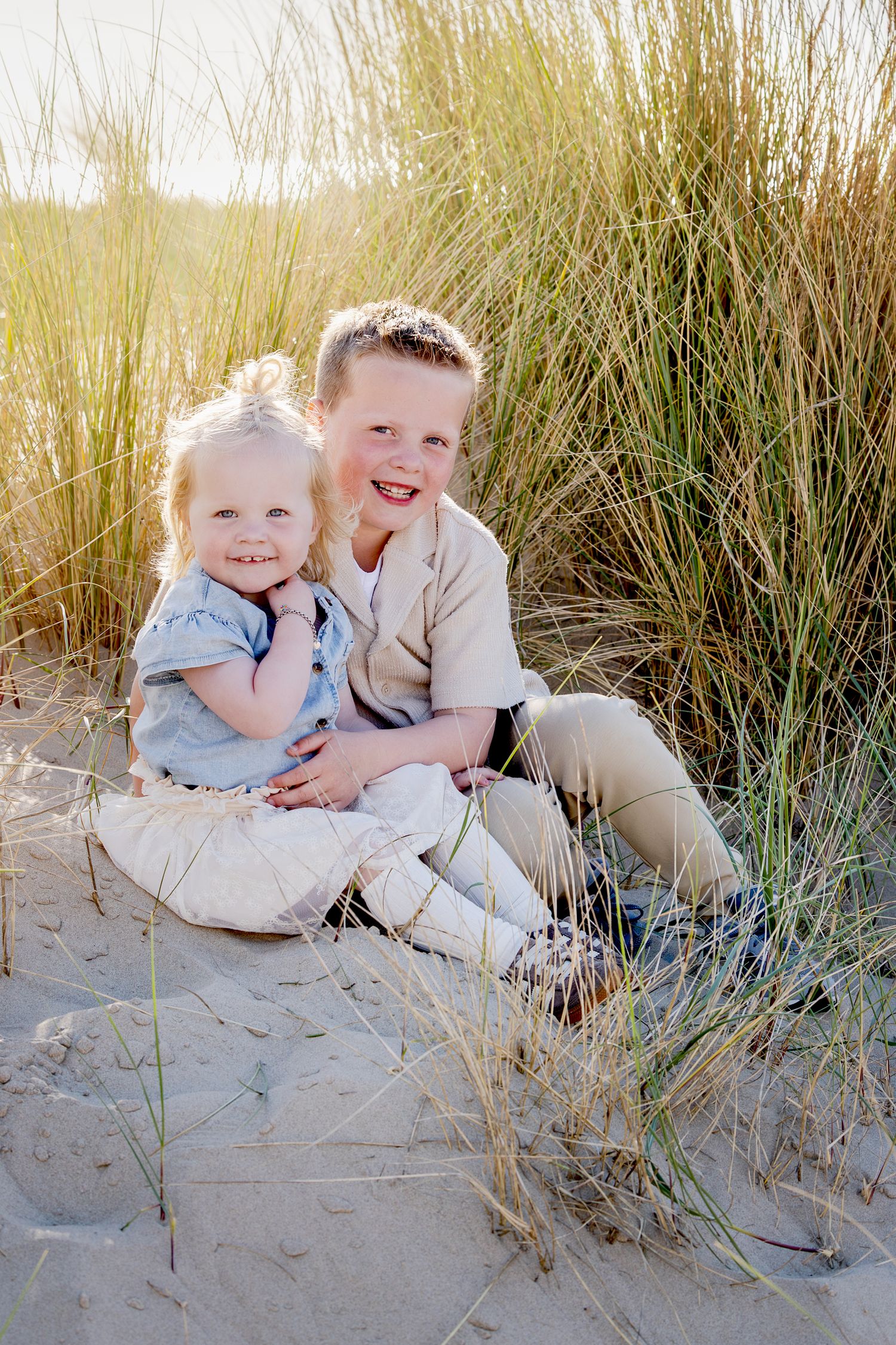 broer (5) en zus (2) zitten knuffelend tussen het hoge gras in de duinen bij ondergaande zon.