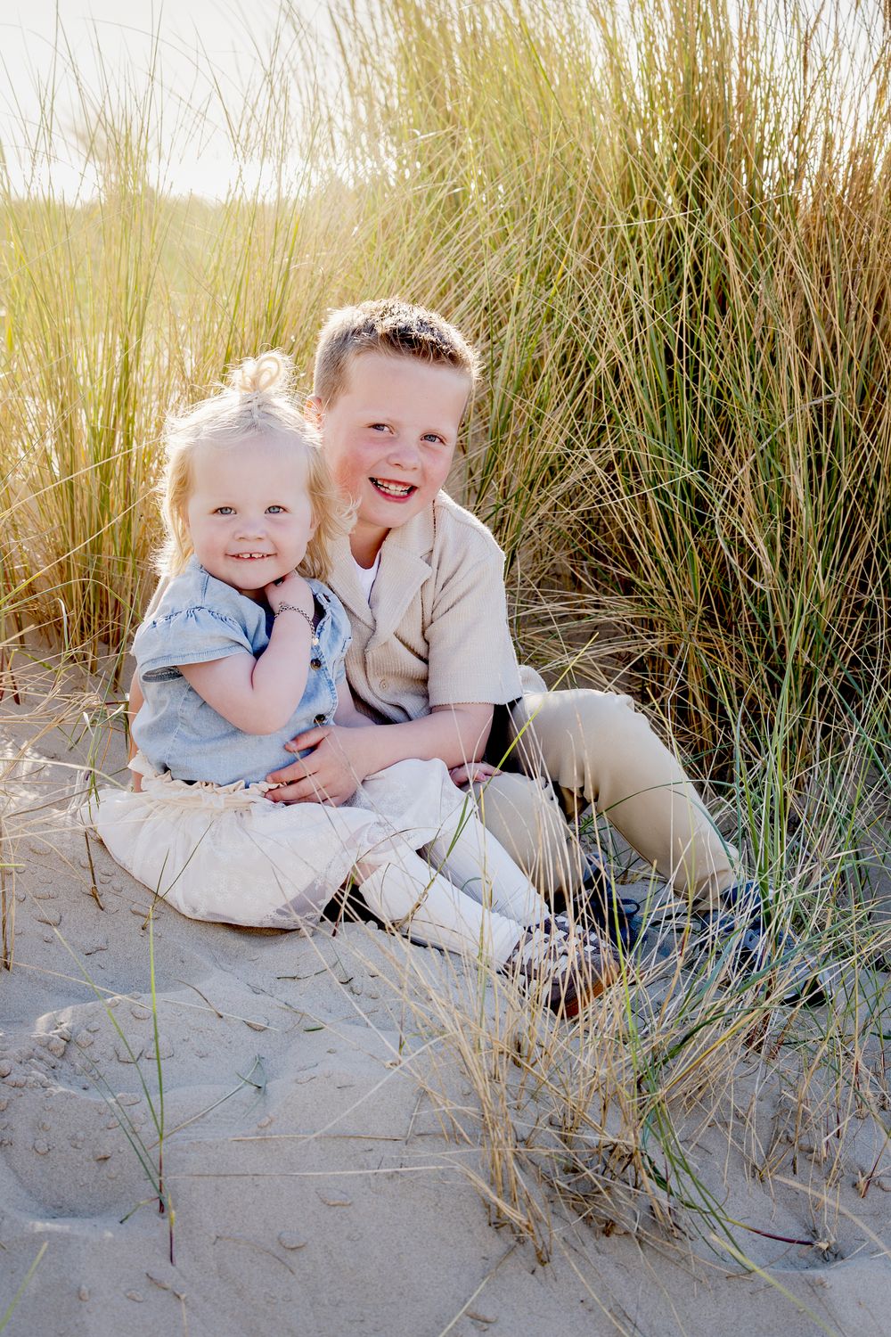 broer (5) en zus (2) zitten knuffelend tussen het hoge gras in de duinen bij ondergaande zon.