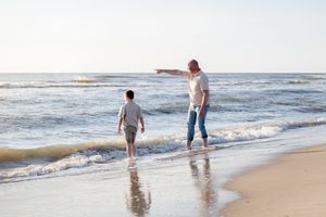 vader en zoon lopen op blote voeten door de branding. vader wijst naar een boot in de verte, terwijl de zon in zee zakt.