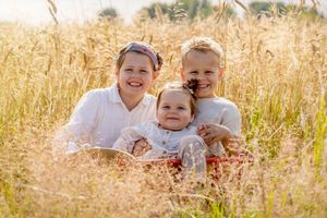 twee zusjes en een broertje. zittend in een kleine houten bolder in het hoge gras met op de achtergrond een graanveld.