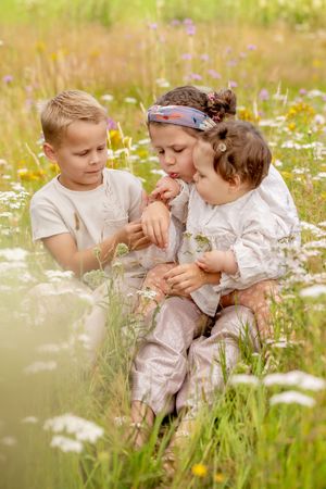 drie jonge kinderen, zittend in een bloemenveld. grote zus heeft een lieveheersbeestje op haar hand die ze wegblaast.
