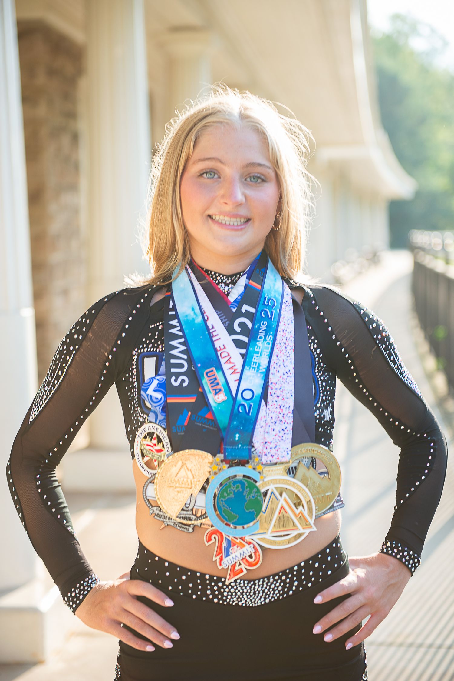 A dancer wearing a black performance dress displays multiple medals and ribbons around their neck.
