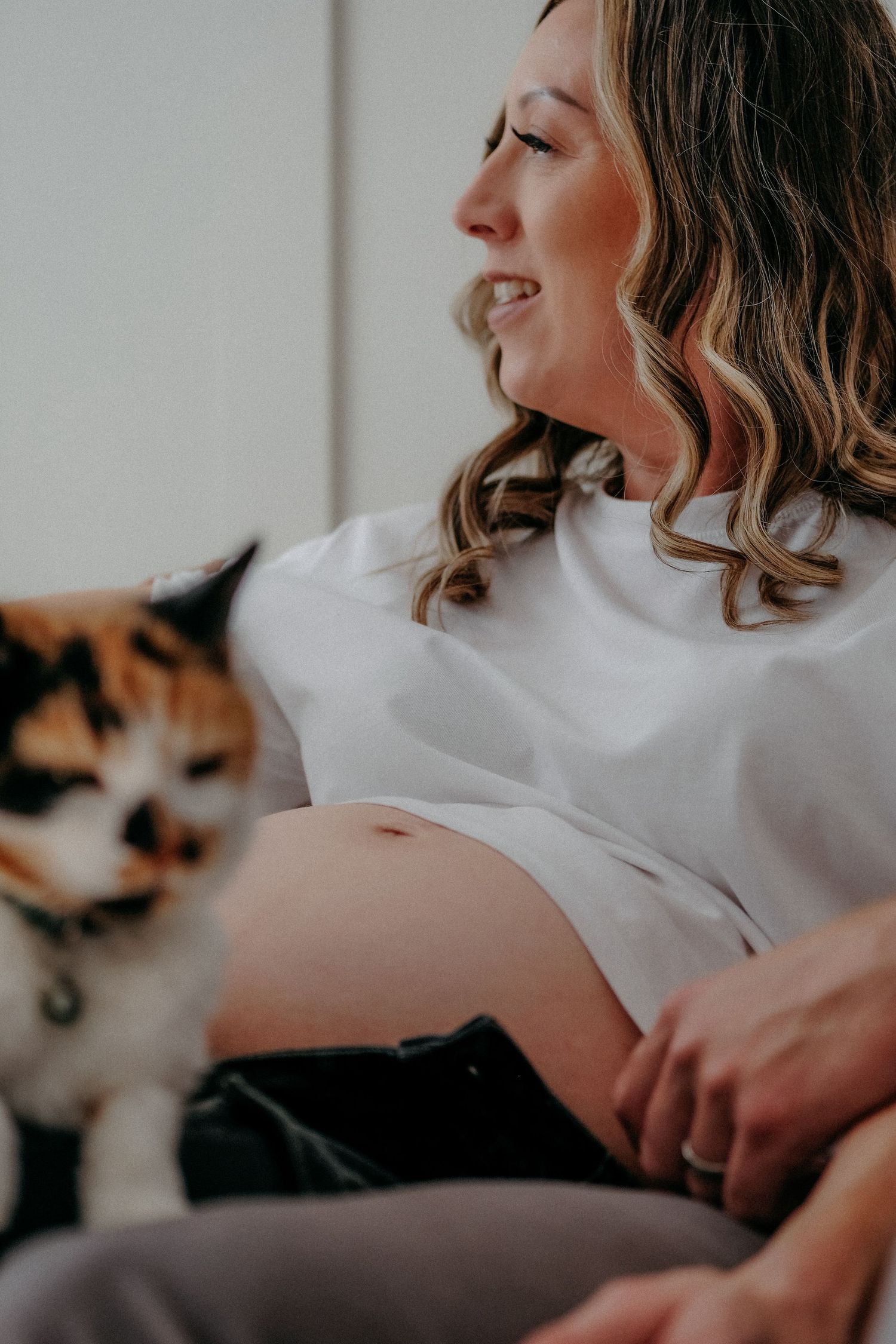 Pregnant mother sitting indoors holding her bump with a pet cat beside her during a relaxed in-home maternity photography session.