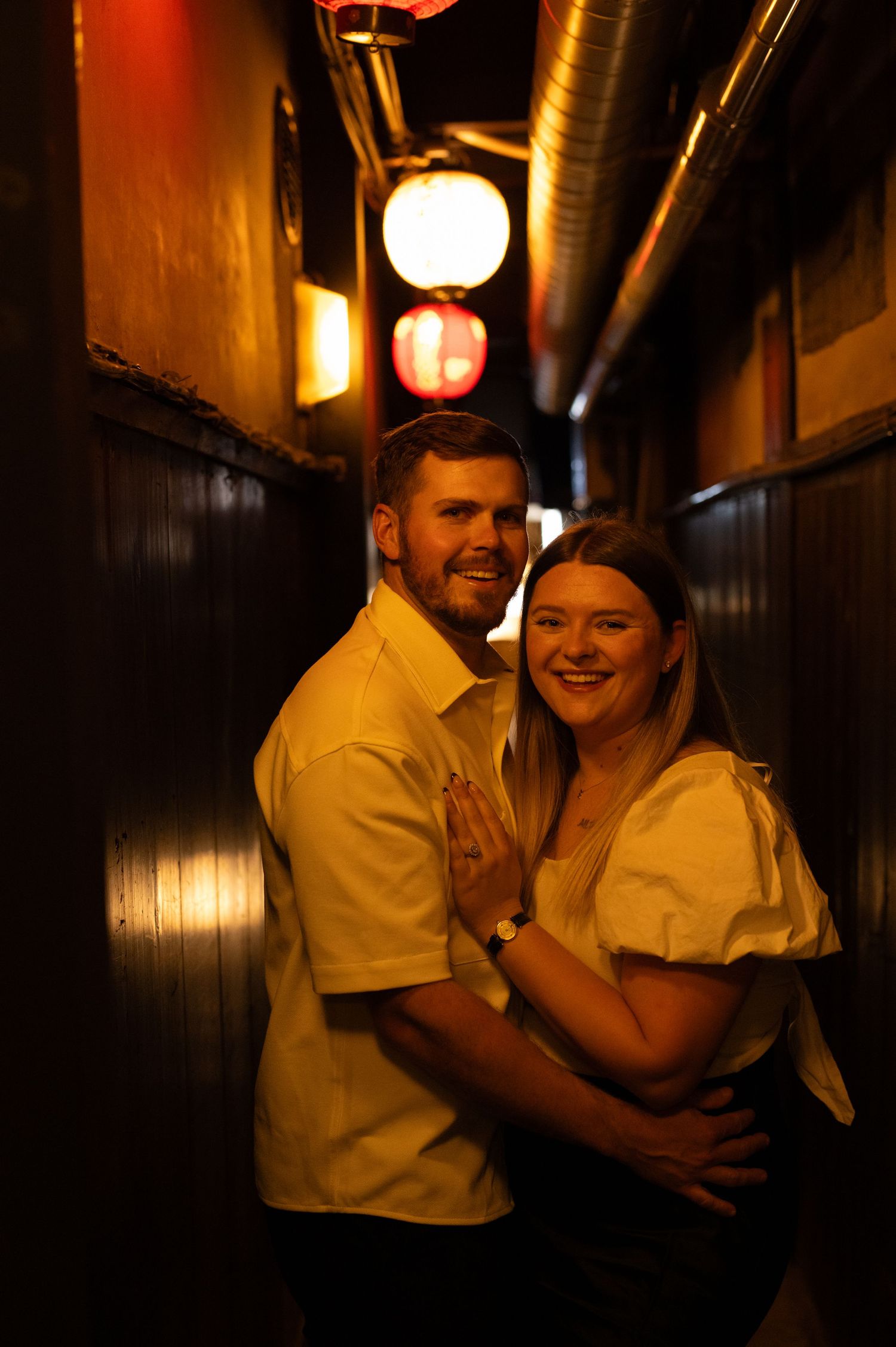 Smiling couple posing together in a narrow lantern-lit Japanese alleyway at night.