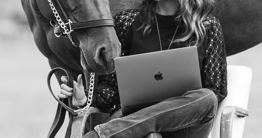 Person sitting on white chair working on MacBook laptop while holding horse reins in black and white photo.