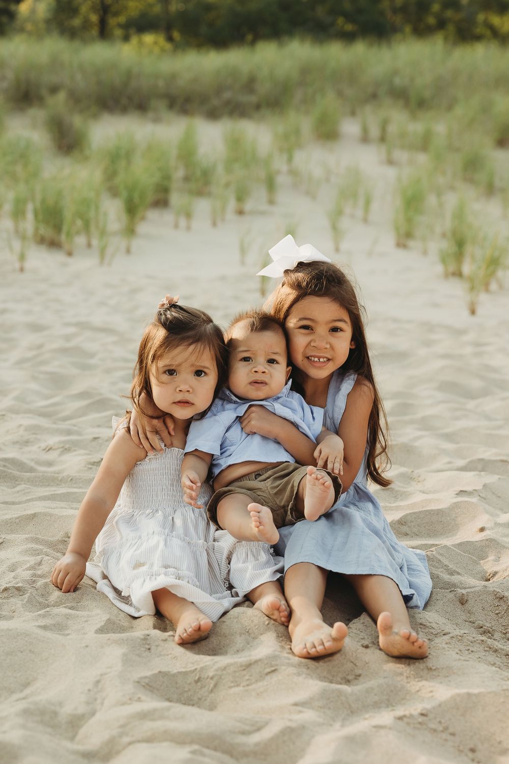Three children in light summer clothing sit together on a sandy beach with beach grass in the background.