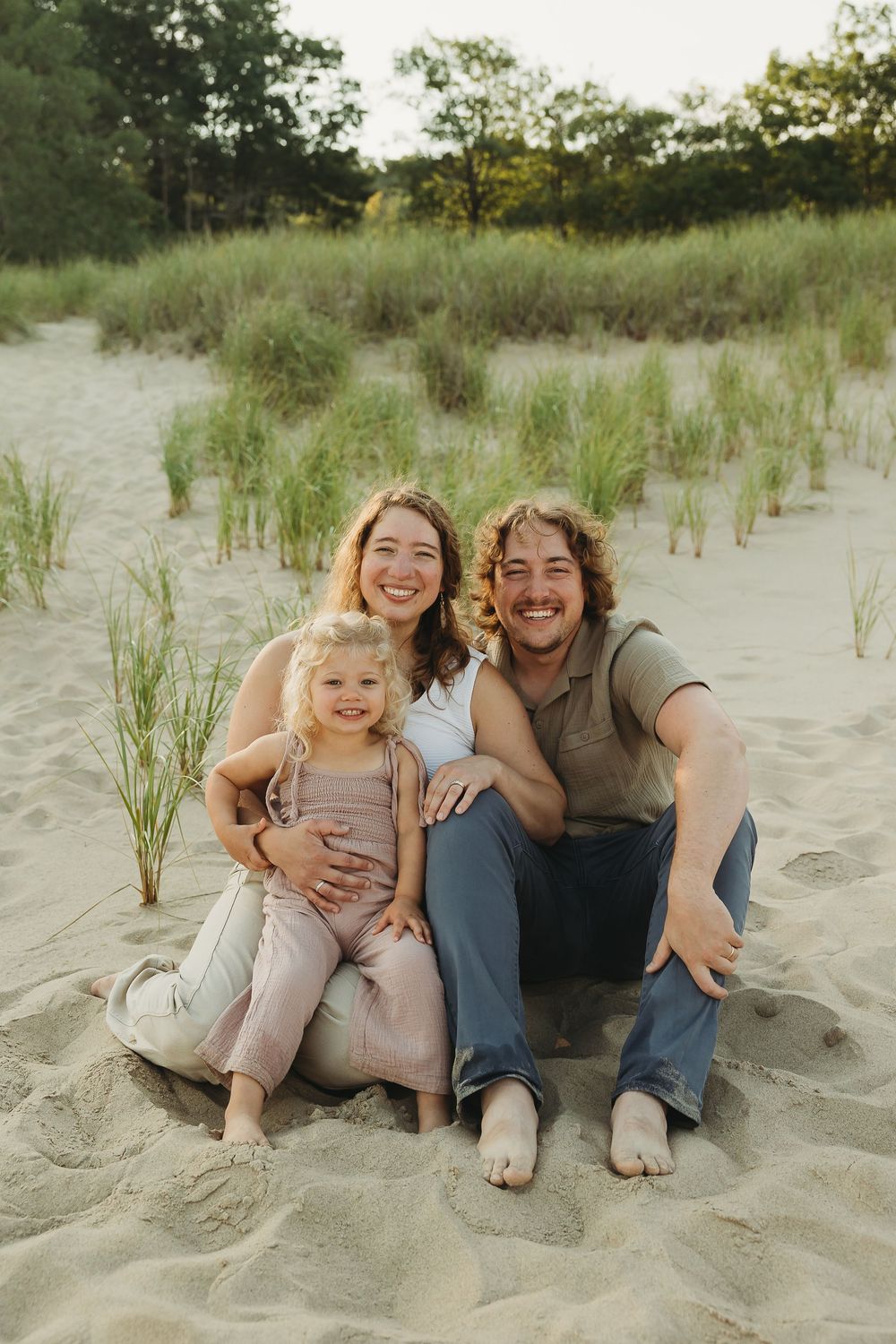 Family sitting together and laughing on sandy beach dunes during golden hour sunset.