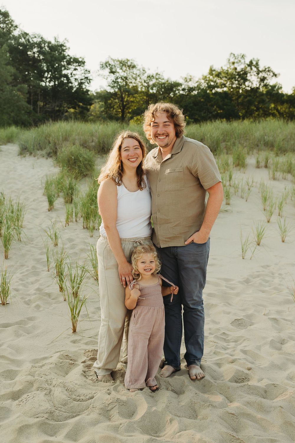 Family wearing neutral colors poses together on sandy beach dunes at sunset.