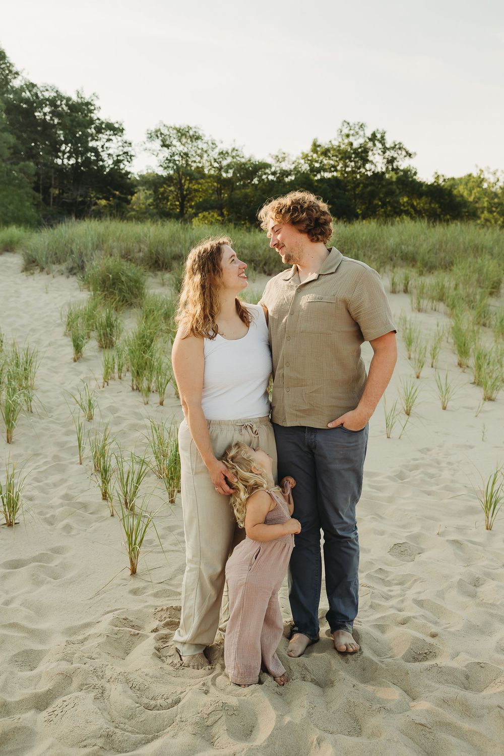 Family stands together on sandy beach with tall grass and trees during golden hour sunset.