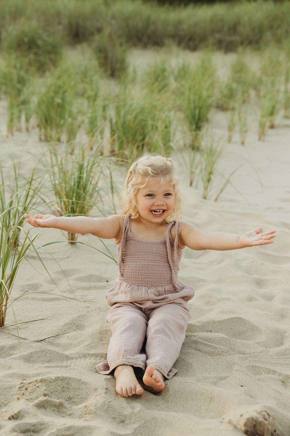 A joyful child sits on sandy beach with arms outstretched among beach grass on a sunny day.