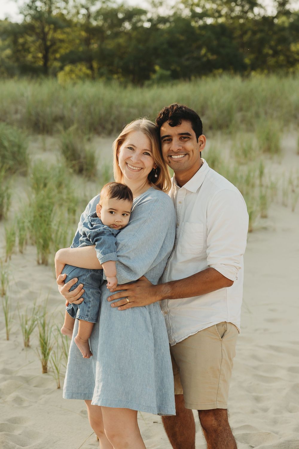 A couple embraces on a sandy beach with tall grass and dunes in the background during a warm summer day.