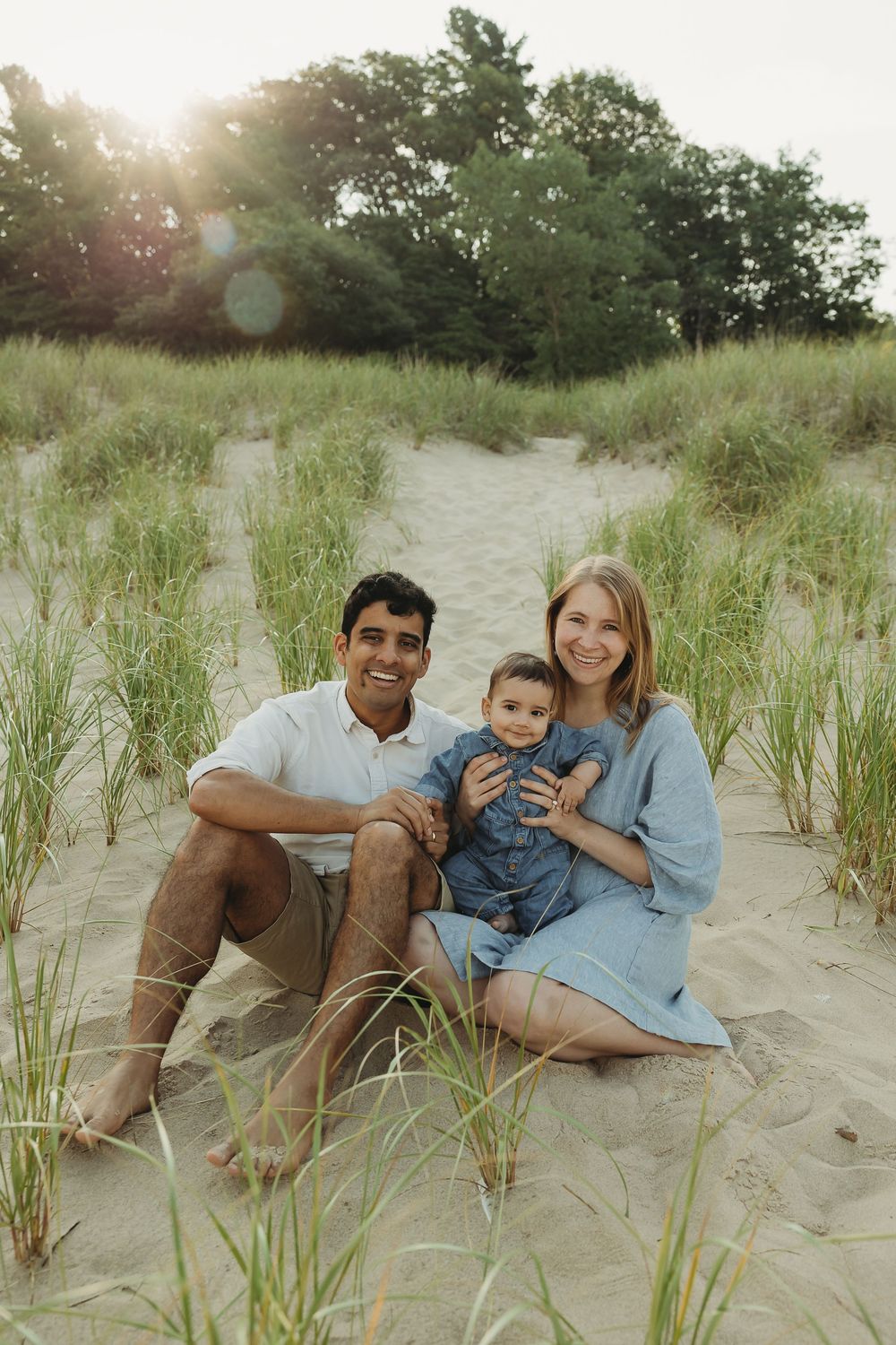 A family sits casually on beach sand surrounded by dune grass during a golden hour photoshoot.