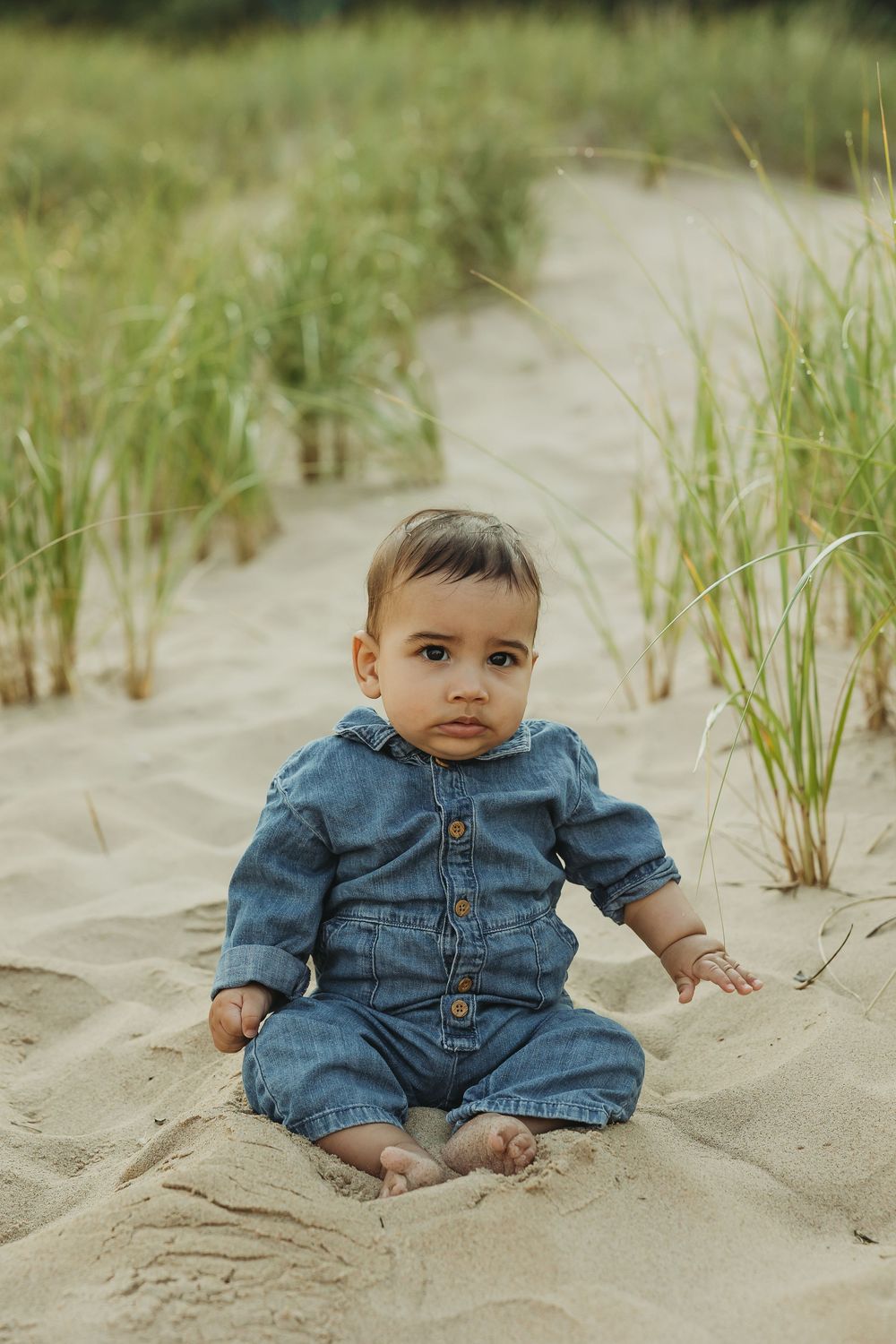 A baby in denim overalls sits on sandy beach dunes with sea grass growing in the background.