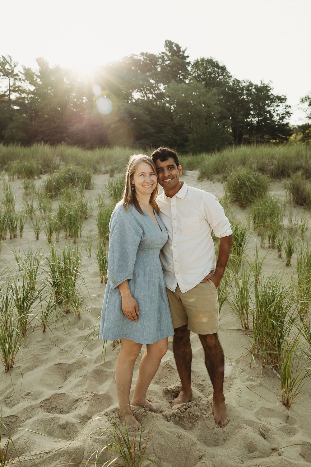 A couple stands together on sandy dunes during golden hour as sunlight filters through trees in the background.