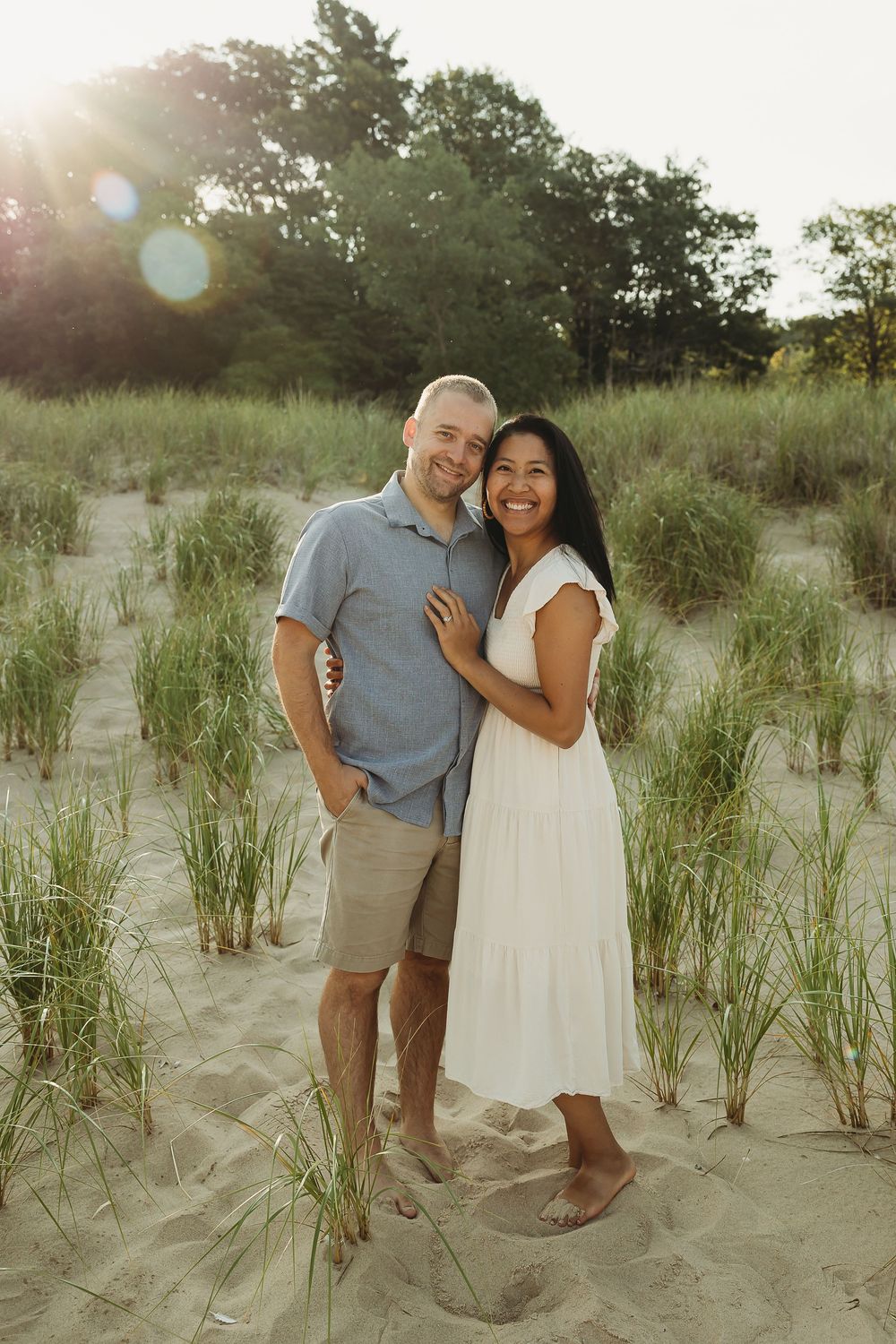 A couple shares joyful moments together on a sandy beach with tall grass and warm sunlight in the background.