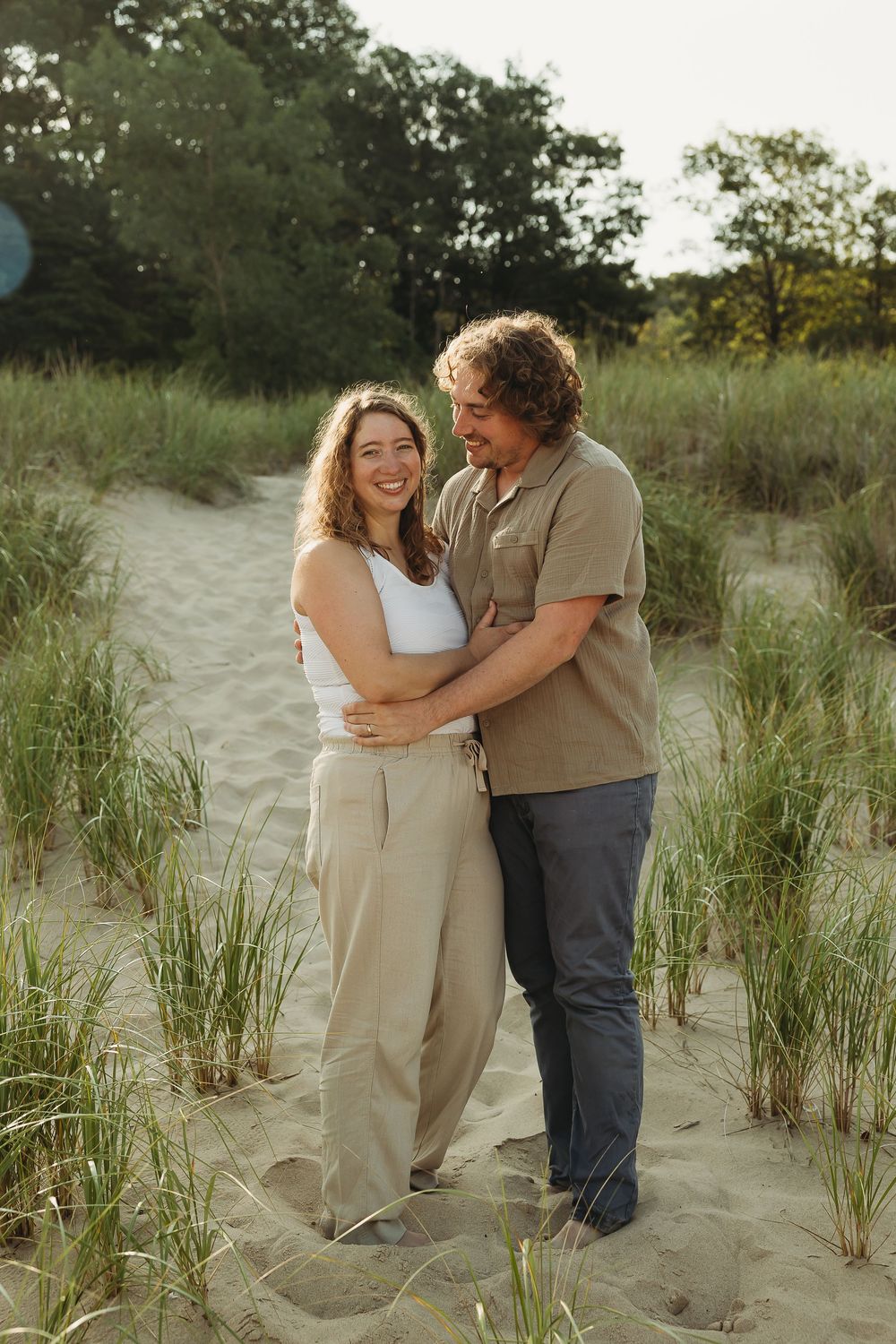 A couple shares tender moments together on a sandy beach surrounded by tall grass during a summer photoshoot.