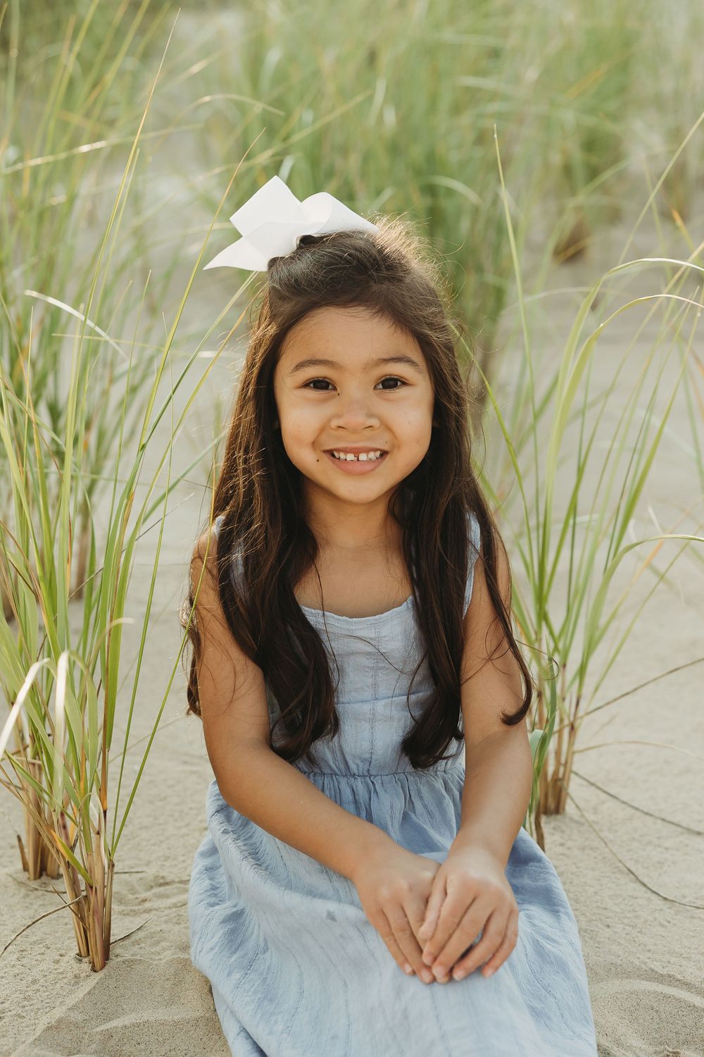 A child in a light blue dress sits among beach grass and sand dunes on a sunny summer day.