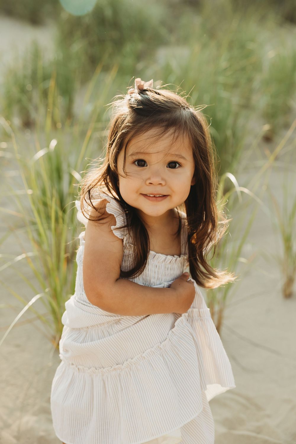 Young child in white ruffled dress plays and smiles on sandy beach dunes with sea grass in background.