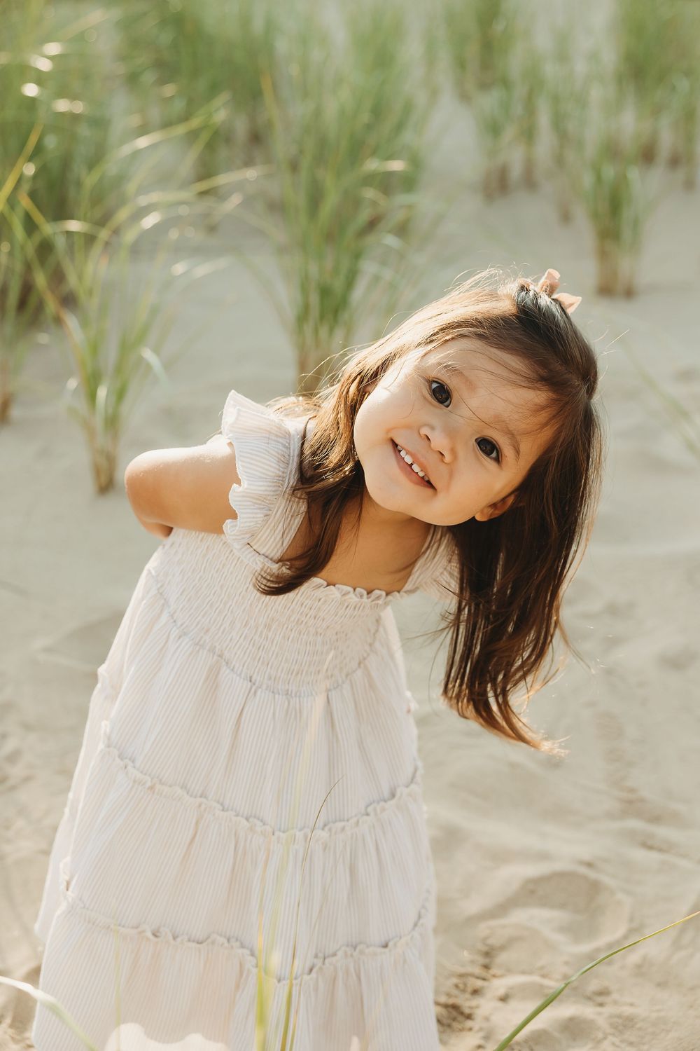 Child in white dress plays joyfully among beach grass and sand dunes on a sunny day.