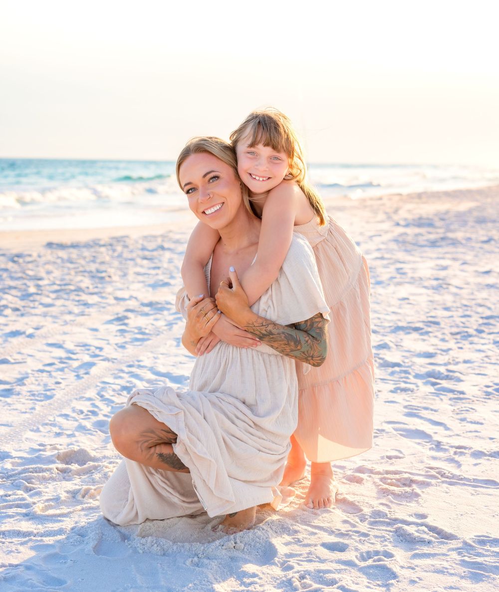 Mother and child share tender moments on a white sandy beach at sunset wearing light colored dresses.