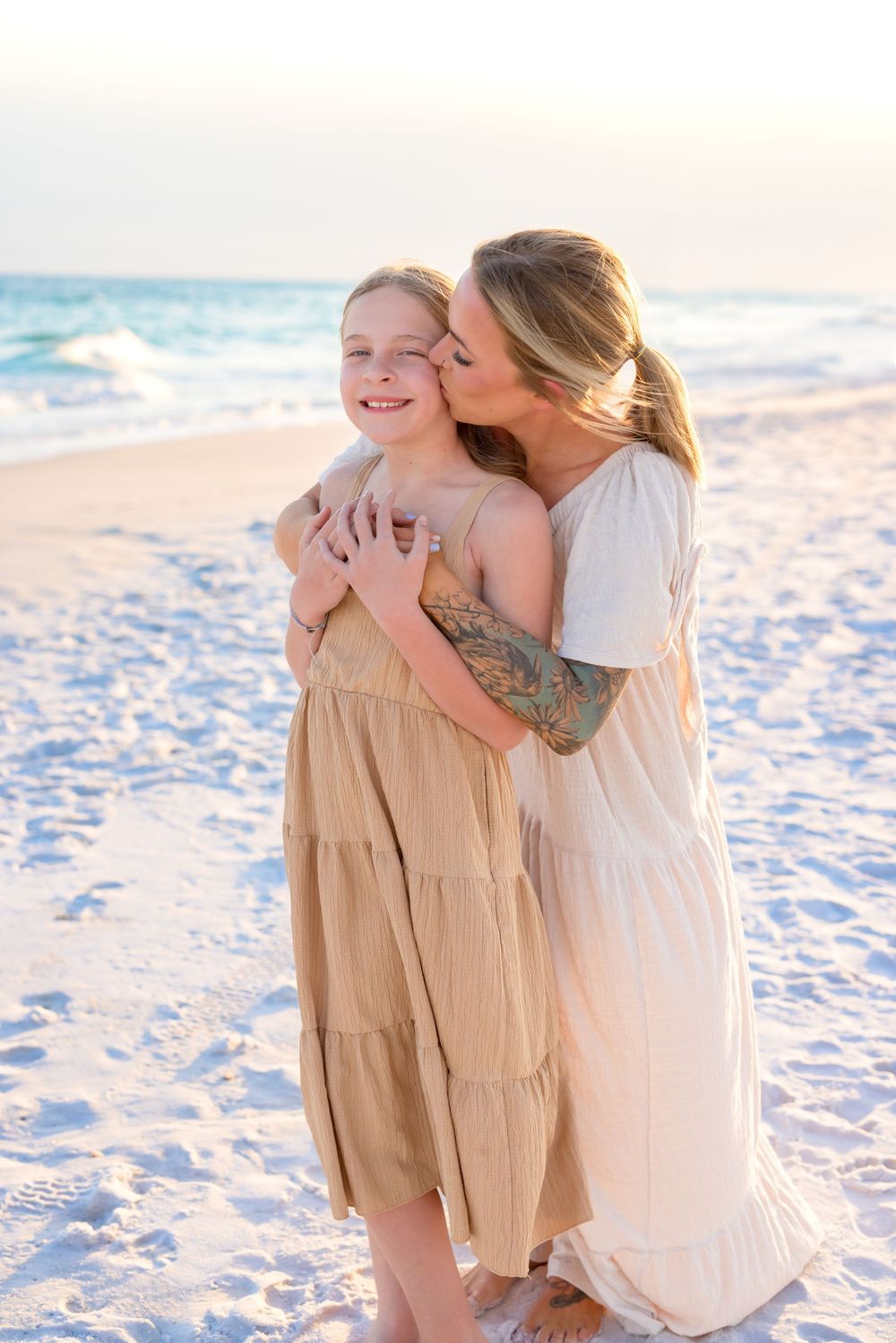 Family embraces on pristine beach with white sand during golden hour wearing neutral colored outfits.