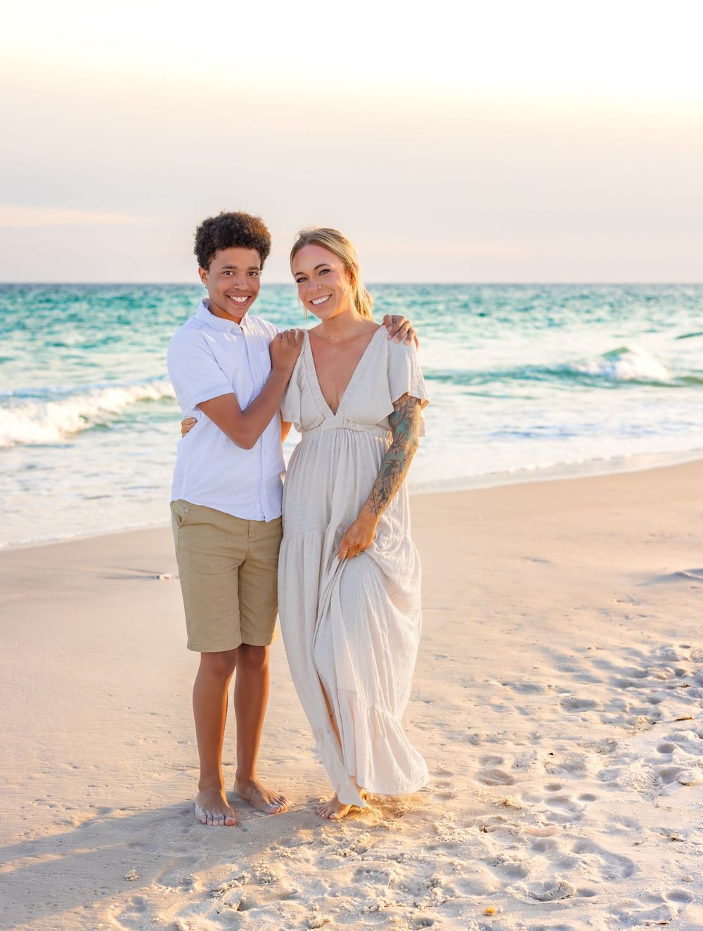 Parent and child walk together on scenic beach at sunset wearing light summer clothing.