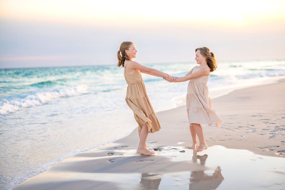 Two people in light colored clothing hold hands while walking along a sandy beach at sunset.