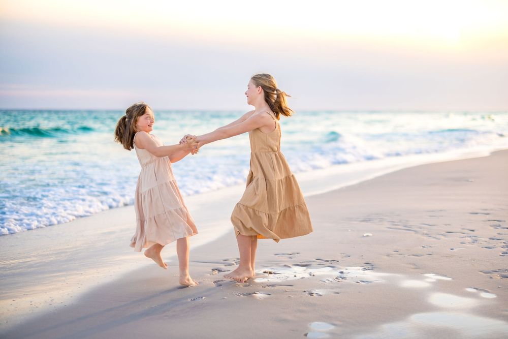 Two people in light flowy dresses walk and dance together along a sunset beach with ocean waves in the background.