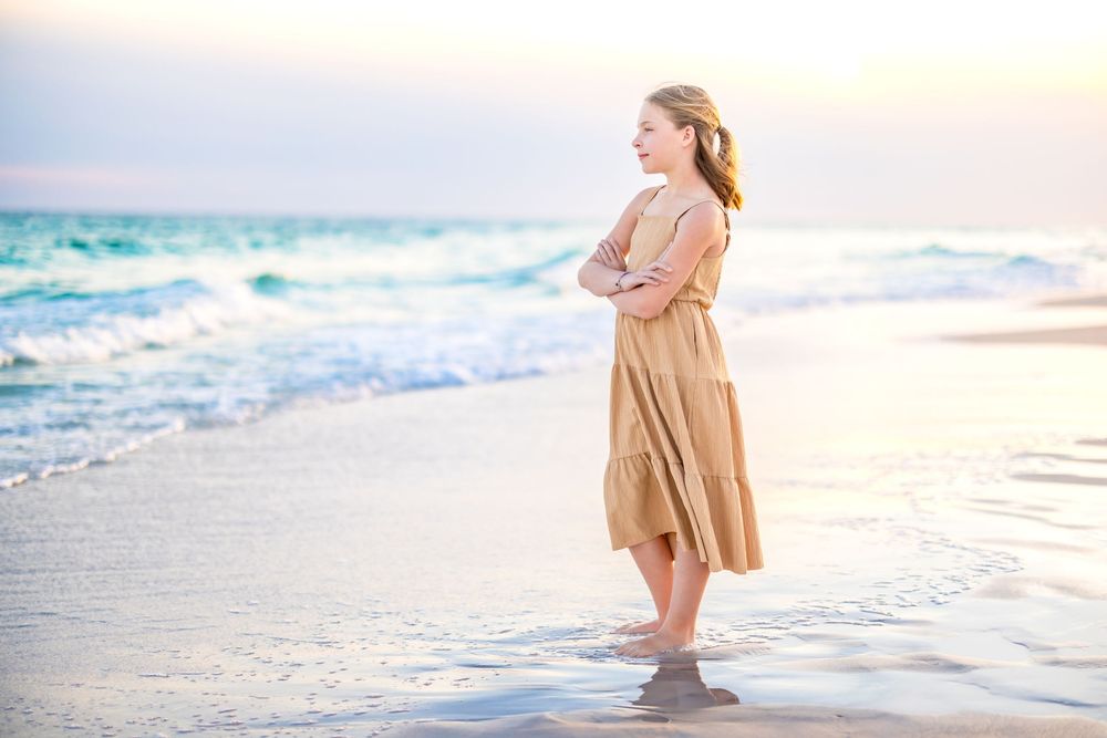 A figure in a flowing beige dress stands barefoot on a beach at sunset with turquoise waves crashing behind them.