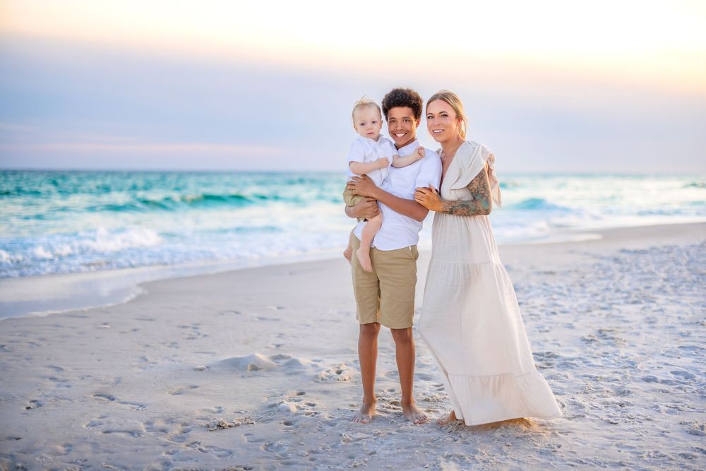 A family dressed in white and beige clothing embraces together on a scenic beach at sunset.
