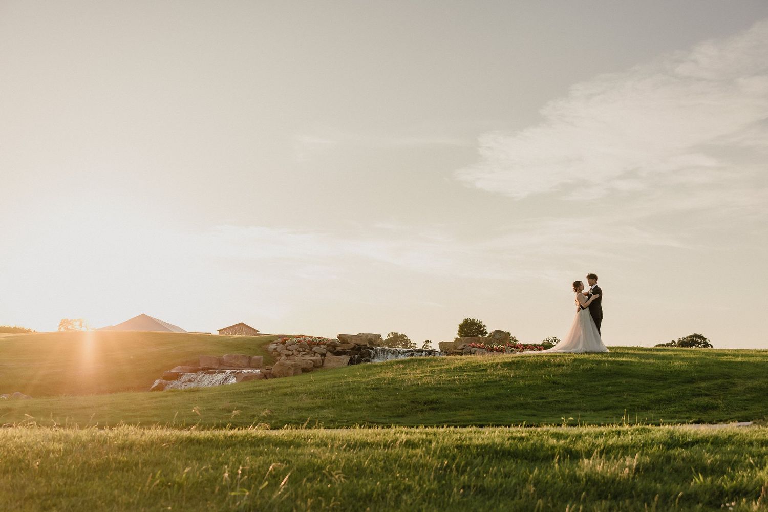 Rolling green hills and fields stretch across a scenic landscape during golden hour with silhouettes visible on the horizon.