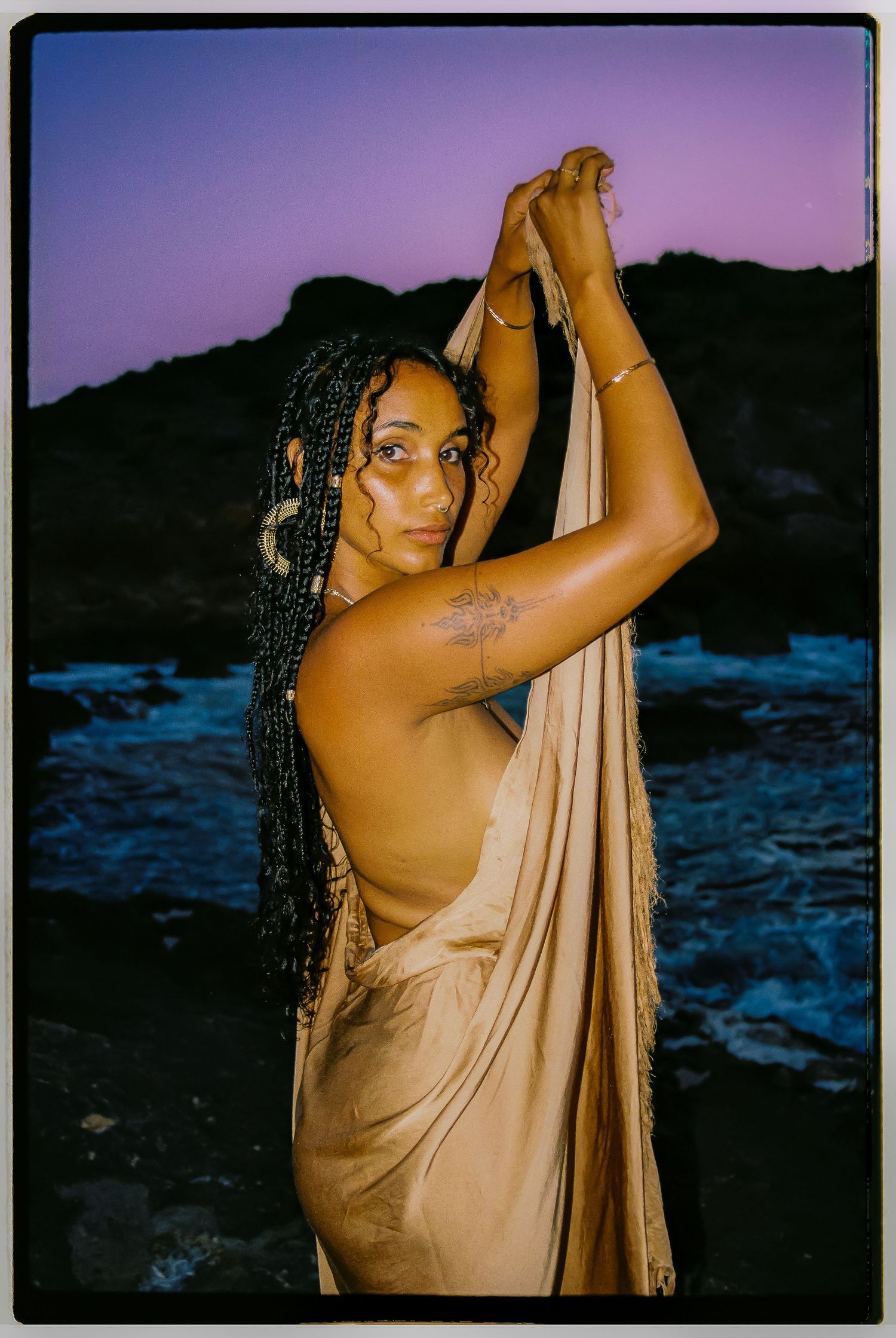 Artistic portrait of a person in flowing beige fabric against a purple sunset sky and rocky coastal landscape.