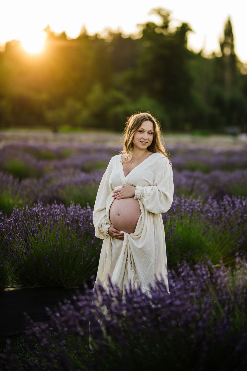 A person in a white dress poses in a blooming lavender field at sunset, cradling their pregnant belly.