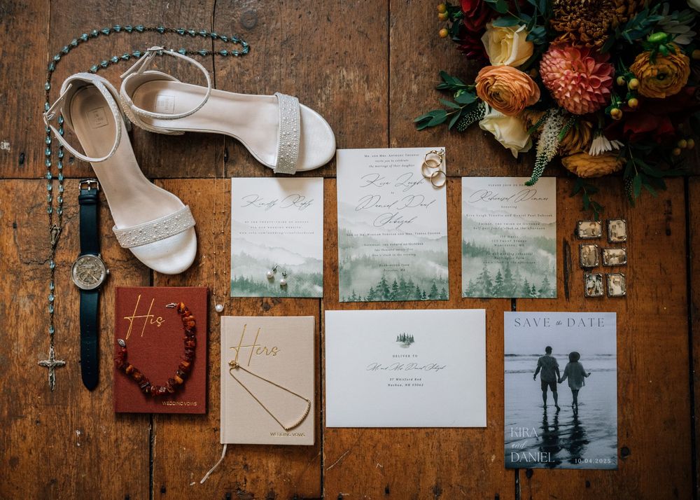 Wedding details flatlay with white heels, invitations, rings, and floral arrangements on rustic wooden surface.