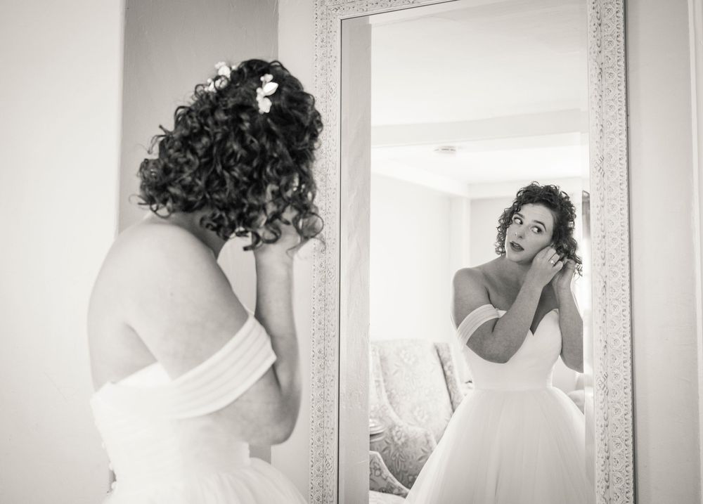 A bride in a white dress looks at her reflection in a mirror during wedding preparations.