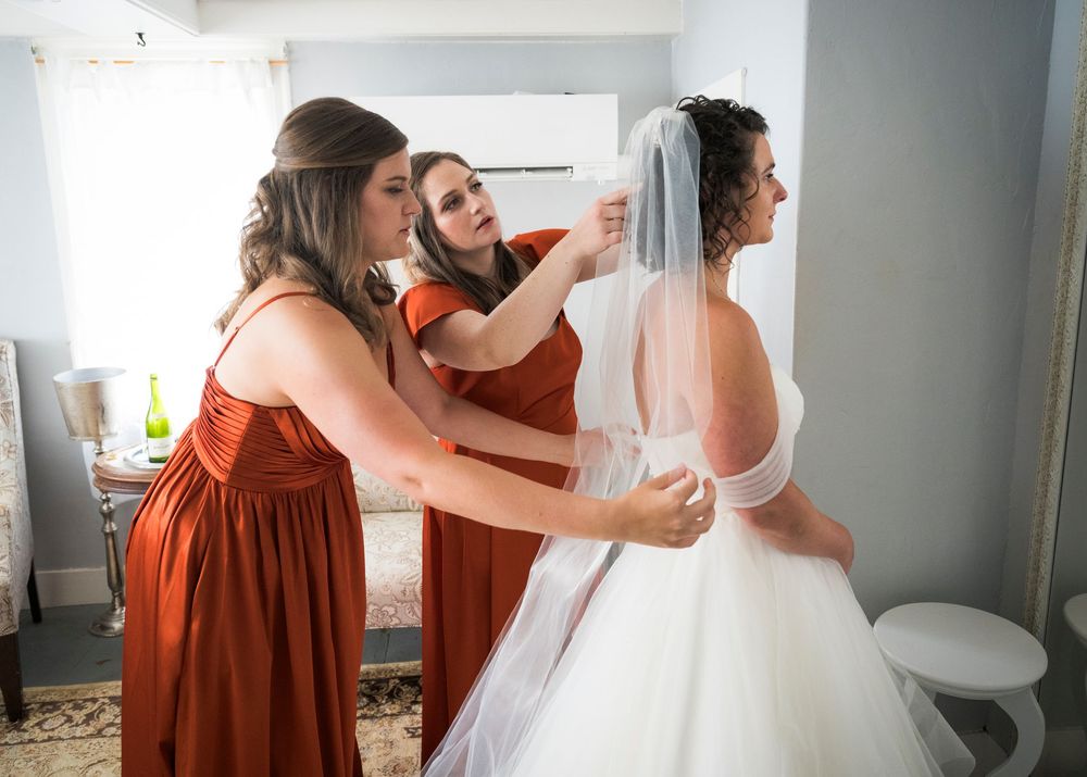 Bride in white wedding dress getting ready with help from bridesmaids wearing rust-colored dresses.