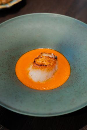 A seared scallop served on bright orange sauce on a textured blue-grey plate.