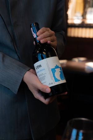 Hands holding blue-labeled wine bottles in a dimly lit restaurant setting.