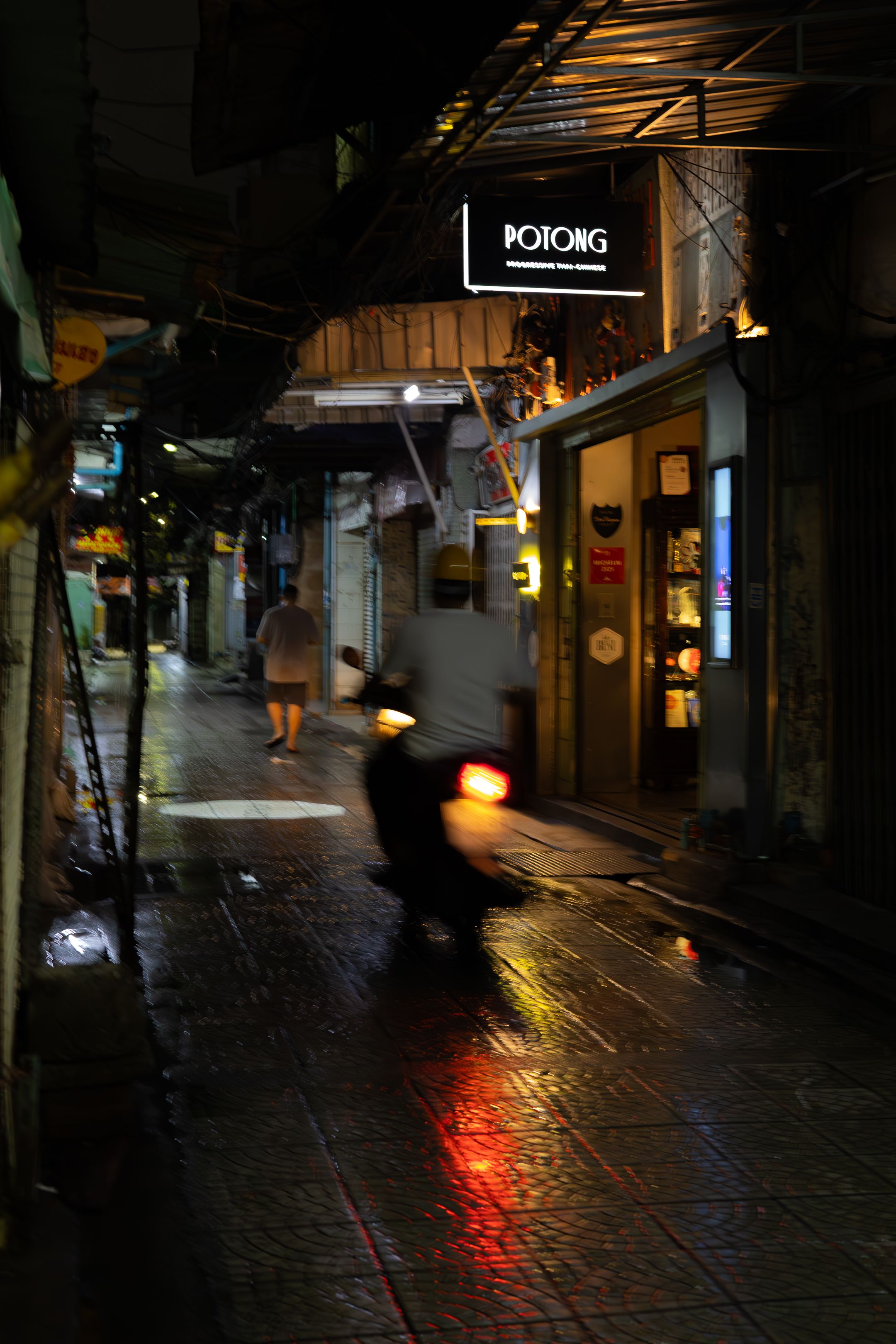 Dimly lit narrow street at night with red lights reflecting off wet pavement and distant storefronts.