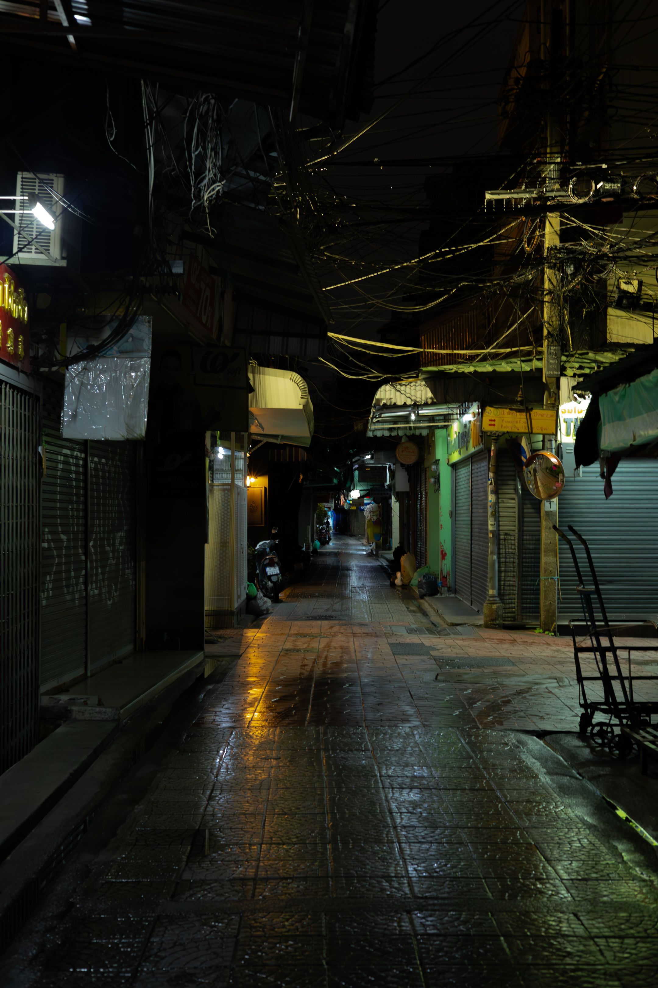 Dark narrow alley in an Asian city with closed shops and glowing lights reflecting off wet pavement at night.