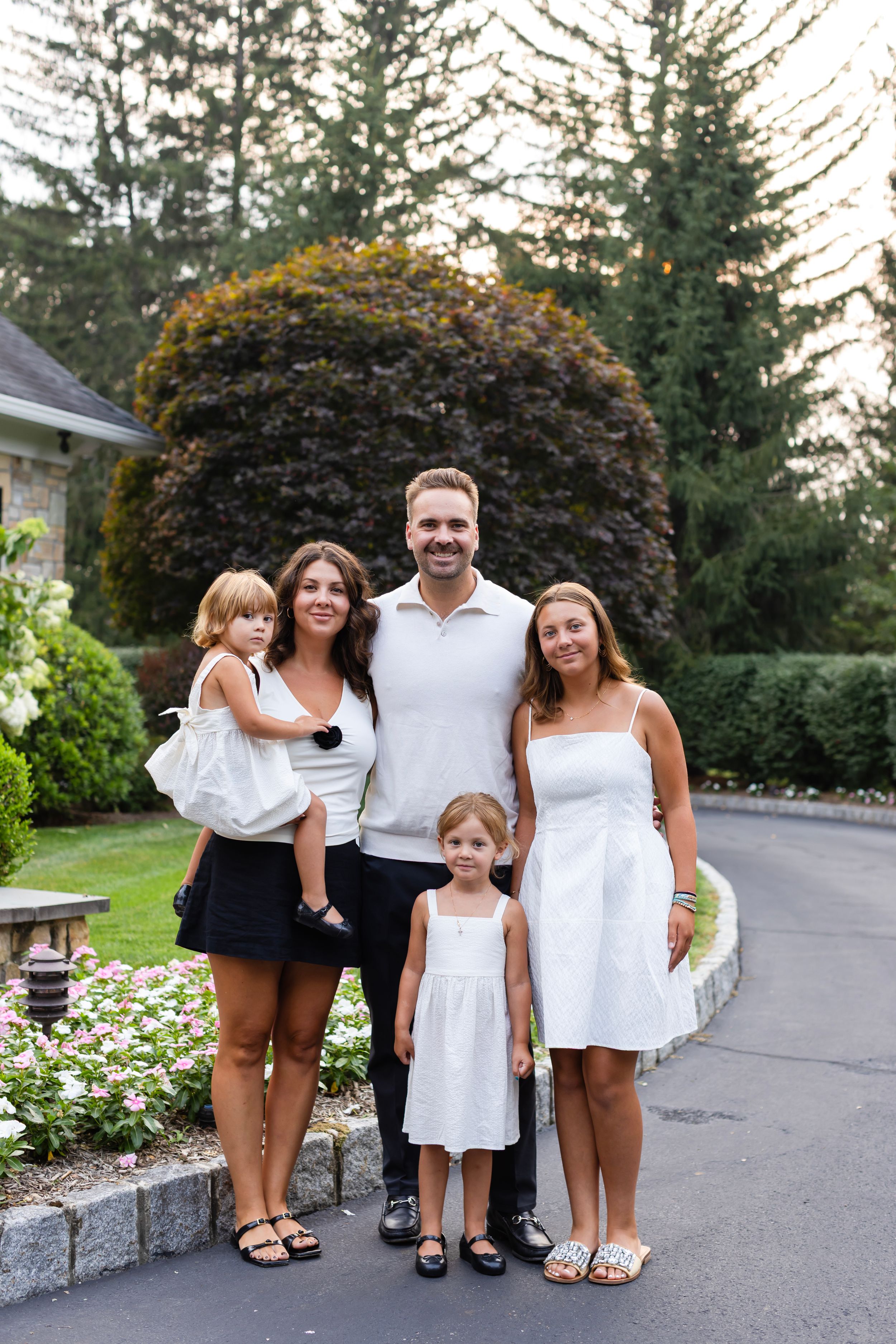 A family wearing coordinated white outfits poses together outdoors on a paved pathway near landscaped gardens.