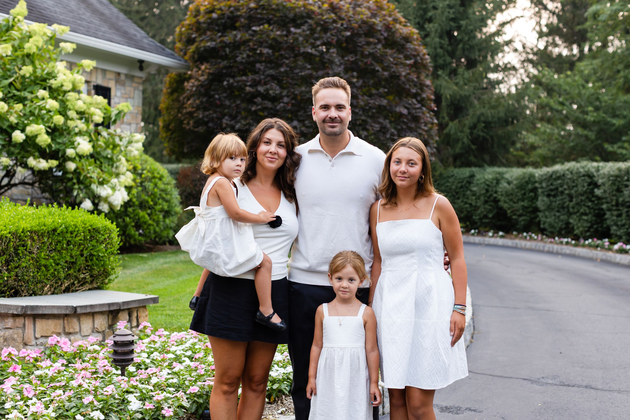 A group poses together outdoors on a driveway with landscaped gardens and greenery in the background.