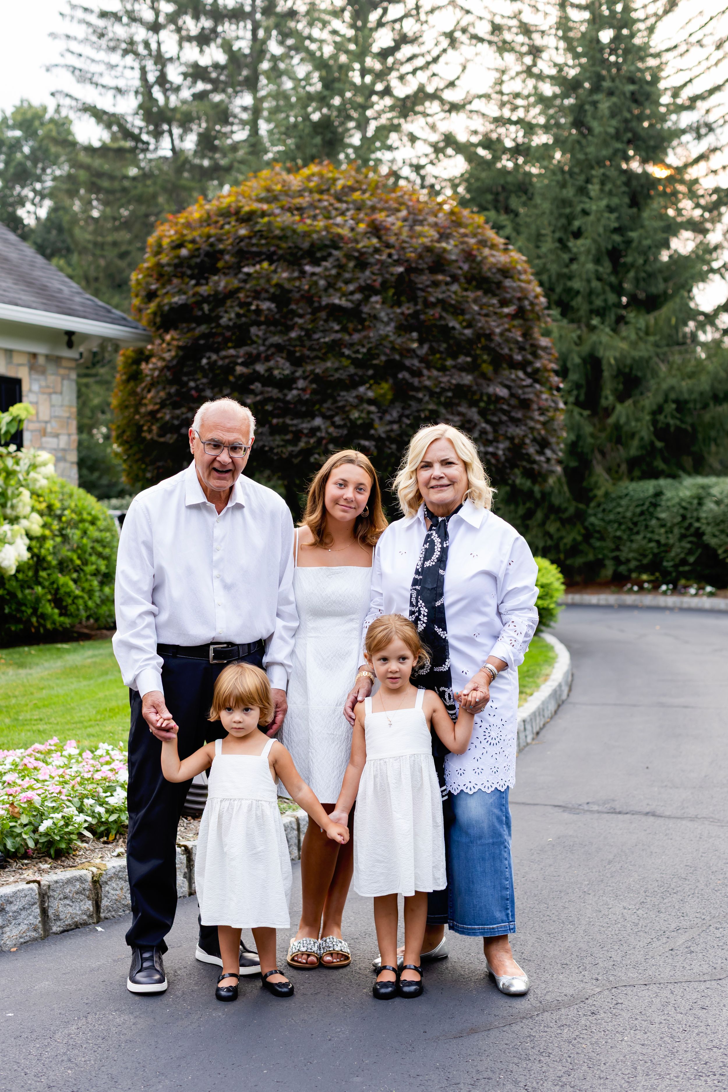 Family members in coordinating white outfits pose together on a paved walkway surrounded by landscaping.
