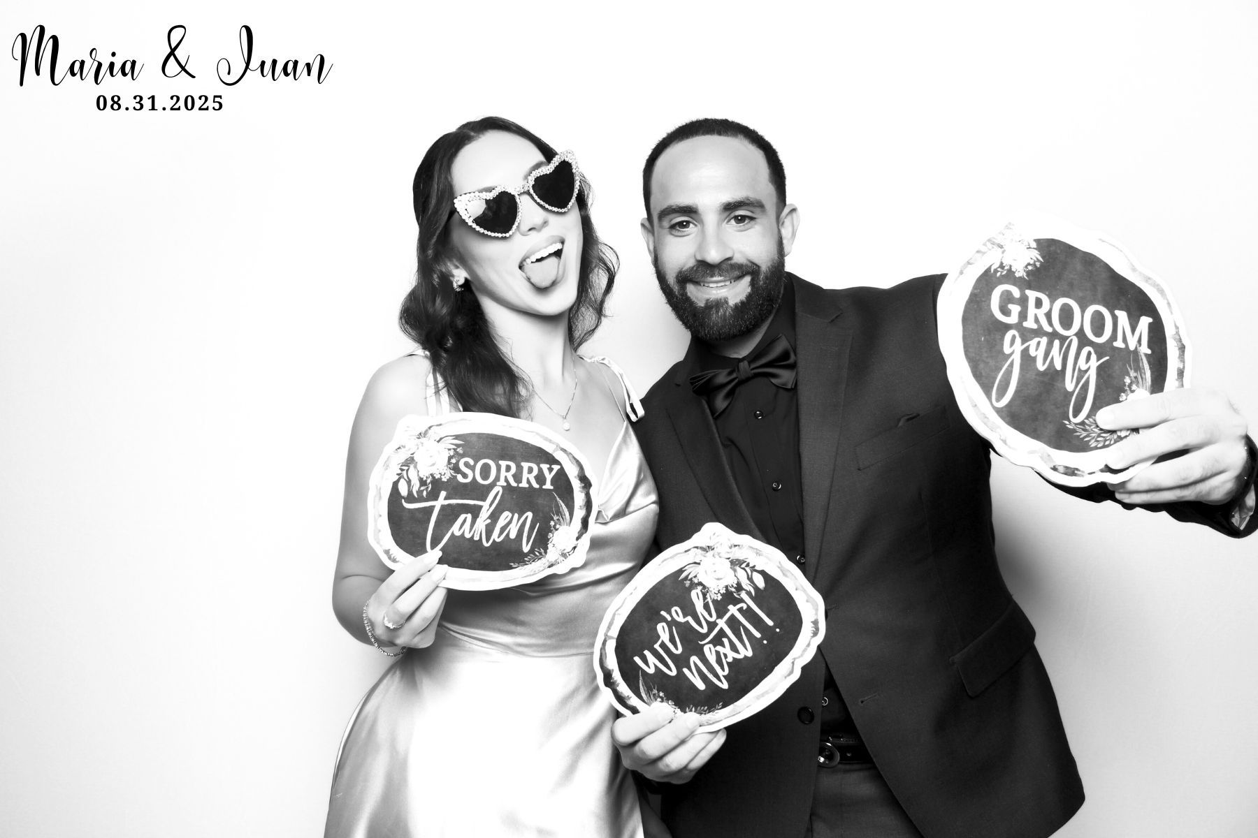 A black and white photo booth image showing a couple posing with fun wedding signs at their celebration.